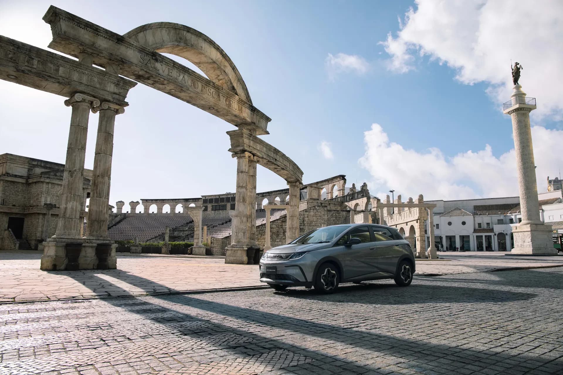BYD Dolphin warna abu-abu di plaza berbatu dengan gapura bergaya Romawi